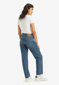 Blue denim straight-leg jeans with a high waist, visible back pockets, and a leather brand patch at the rear. Paired with a white T-shirt.