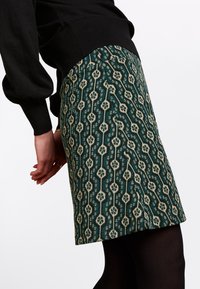 Grüner gemusterter Rock mit strukturiertem Design und cremefarbenen sowie schwarzen Akzenten, knielang, kombiniert mit schwarzen Strumpfhosen und einem schwarzen Oberteil.