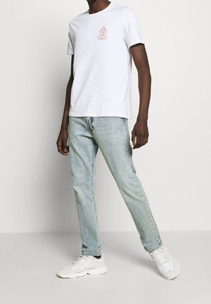 T-shirt en coton blanc avec un motif de flamme rouge, associé à un jean bleu clair et des baskets blanches, présentant une coupe décontractée et un design minimaliste.