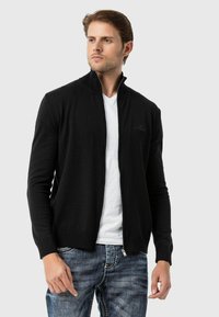 Schwarze Zip-Up-Strickjacke aus gestricktem Stoff, mit hohem Kragen, gerippten Bündchen und Saum sowie einem dezenten Logo auf der Brust.