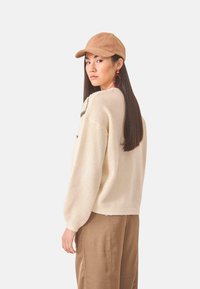 Suéter de crema con mangas globo, gorra beige y grandes pendientes rojos. Textura suave, corte holgado y detalle de diseño sutil en el escote.