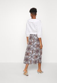 Femme aux cheveux courts et bouclés portant un chemisier blanc à manches bouffantes, une jupe midi à motif floral et des talons argentés ouverts à l'arrière, debout de dos.