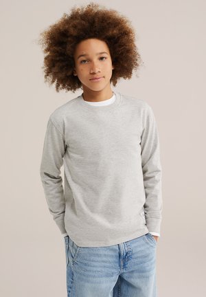 Adolescent aux cheveux bouclés portant un sweat-shirt gris clair et un jean bleu se tient devant un fond beige uni.