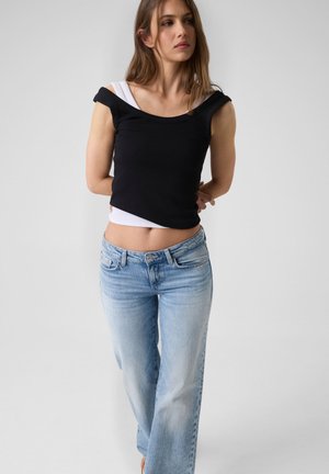 Jeune femme portant des hauts superposés noirs et blancs avec un jean bleu clair, debout devant un fond clair uni.