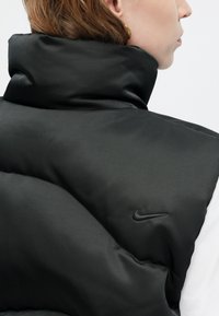 Černá péřová vesta s vysokým límcem, vyznačující se hladkým povrchem a diskrétním černým logem Nike na rameni.