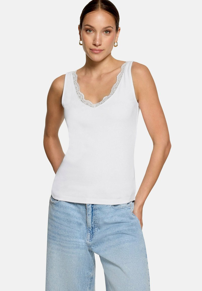Femme portant un haut blanc sans manches avec bordure en dentelle et un jean bleu clair, debout les mains dans les poches devant un fond uni.