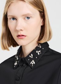 Schwarzes Hemd mit einem Knopfkragen, der mit klaren Edelsteinen in Blumenformen und verstreuten Designs verziert ist. Glatte Textur, figurbetontes Design.