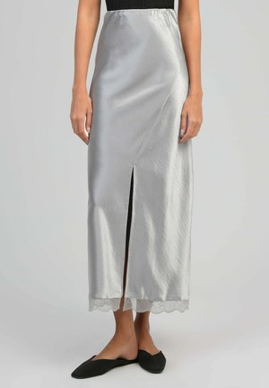 Jupe midi en satin argenté avec fente frontale et ourlet en dentelle, portée avec des ballerines noires pointues sur une personne debout devant un fond uni.