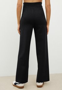 Pantaloni negri cu croială largă, dintr-o țesătură netedă și cu talie înaltă. Purtați cu adidași albi cu accente bleumarin. Vedere din spate.