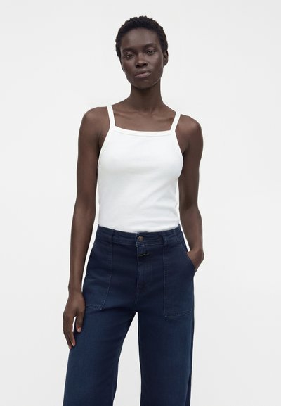 Jeune femme noire debout, portant un haut blanc sans manches et un pantalon bleu foncé taille haute, une main dans la poche, expression neutre.