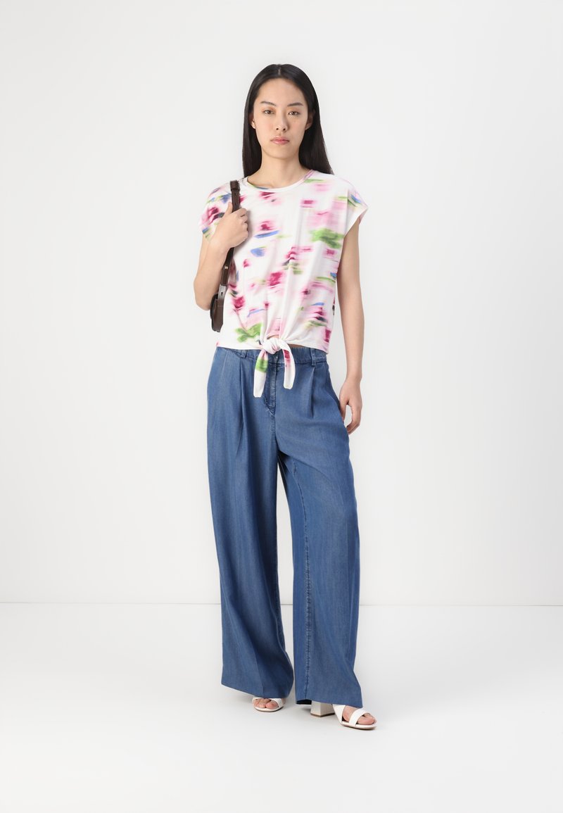 Bluse mit floralem Druck in Weiß, kurzen Ärmeln und geknoteter Vorderseite, kombiniert mit weit geschnittenen blauen Denim-Hosen und weißen, high-heeled Sandalen.
