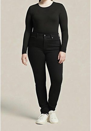 Person trägt schwarzen langärmeligen Oberteil und schwarze hoch taillierte Hose, steht mit gekreuzten Beinen, trägt weiße Sneaker vor neutralem Hintergrund.