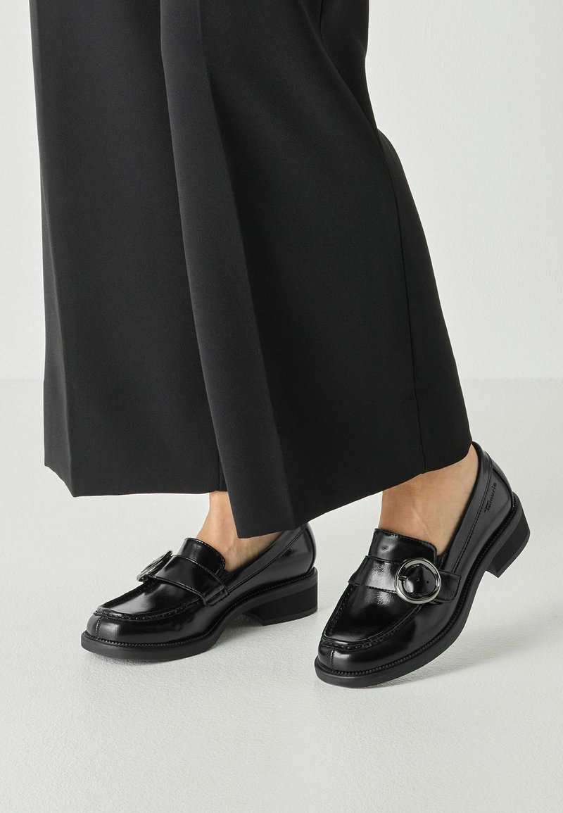 Mocassins en cuir noir avec un détail de boucle ronde portés avec un pantalon noir à jambes larges sur un fond de sol clair.
