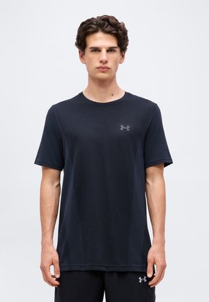 Jonge man met kort golvend haar draagt een effen zwarte Under Armour T-shirt en zwarte shorts, staand tegen een lichtgrijze achtergrond.