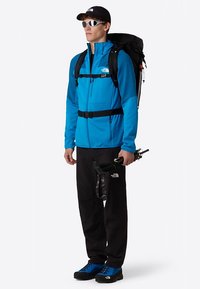 Modrá fleecová bunda se zipem, černé kalhoty, modré turistické boty a černý batoh. Doplňky zahrnují sluneční brýle a turistické hole.