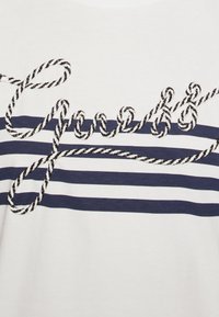 Bílý tričko se námořnickými vodorovnými pruhy, s výšivkou ve tvaru lana přes přední část, vytvářející texturovaný kontrast.