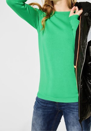 Maglione verde a maniche lunghe in materiale a costine, abbinato a jeans blu, mentre tiene una giacca nera effetto piumino con accenti arancioni.