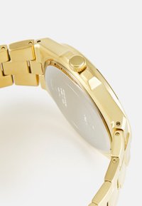 Montre en acier inoxydable doré avec un bracelet à maillons, un boîtier angulaire et une couronne texturée. Présente une surface arrière lisse et réfléchissante.