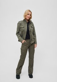 Olive green military-style jacket with four pockets, paired with matching cargo pants. Black shirt underneath, and black footwear.