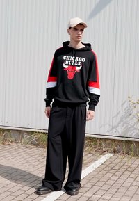 Felpa nera con dettagli rossi e bianchi, che presenta il logo "Chicago Bulls". Pantaloni larghi neri e scarpe nere; modello che indossa un cappellino beige.