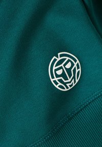 Tessuto turchese con una superficie liscia presenta un logo circolare bianco con un design di volto astratto. Cuciture visibili lungo il bordo.