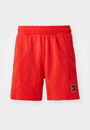 Red cotton shorts with an elastic waistband, side pockets, and a small multicolored logo patch on the left leg.