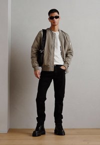 Chaqueta bomber gris, camiseta blanca, jeans negros y botas negras robustas. Unas gafas de sol reflectantes y un bolso negro de hombro completan el atuendo.