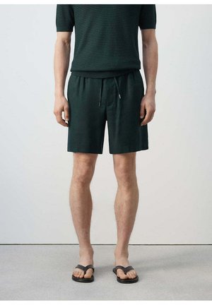 Hombre con camiseta de punto de manga corta verde oscuro, pantalones cortos a juego con cordón y chanclas negras, de pie frente a un fondo claro y liso.