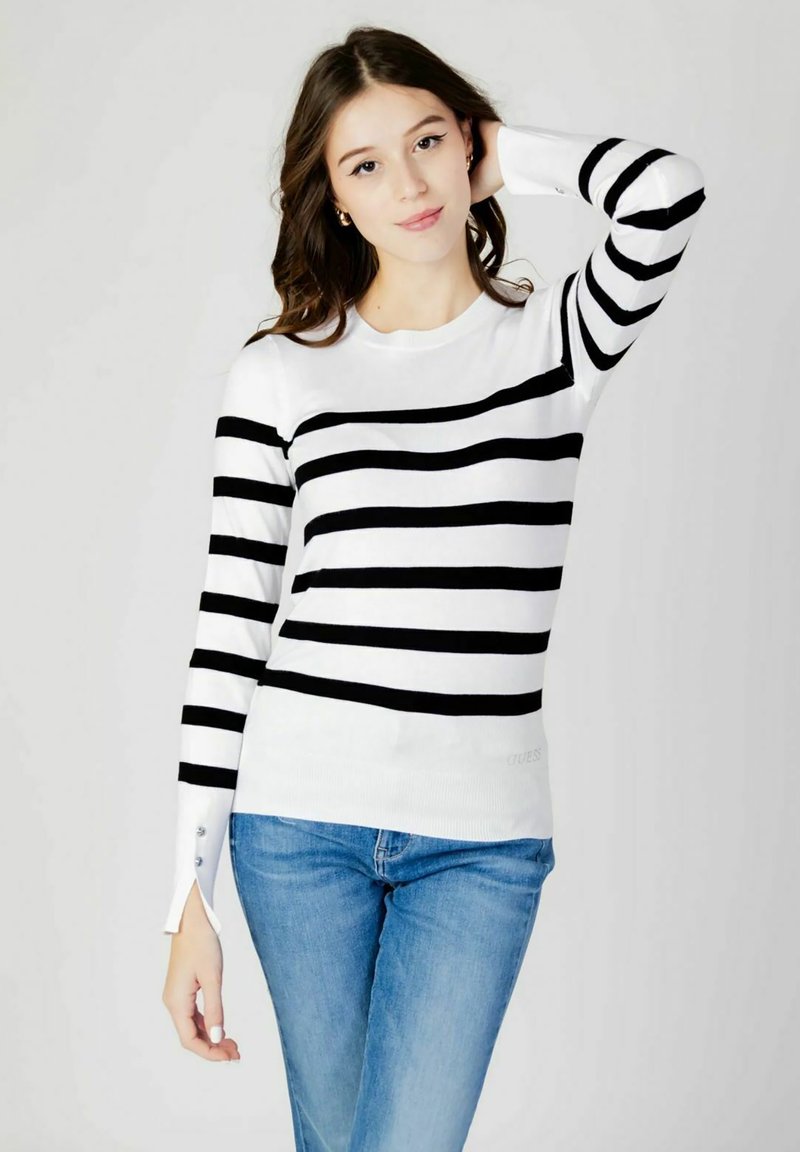Maglione a righe bianche e nere con maniche lunghe, scollo rotondo e dettagli decorativi con bottoni sui polsini. In vista un paio di jeans blu.