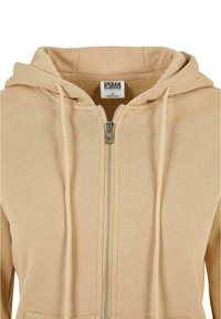 Beige zip-up hettegenser med snøring, med sentral glidelås og frontlommer. Laget av et mykt, strukturert stoff.