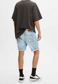 T-shirt noire surdimensionnée, shorts en denim bleu clair, ourlets roulés, patch en cuir marron, chaussettes noires, et baskets noir et blanc.
