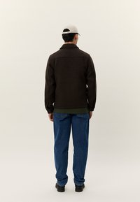 Ruda vilnos mišinio striukė su apykakle, nugaros yoke ir užsegamais rankogaliais, derinama su mėlynais džinsais ir šviesia kepure.