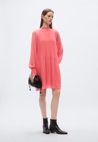 Vestito corallo plissettato con maniche lunghe, collo alto e silhouette fluida. Abbinato a una borsa nera e stivaletti alla caviglia. Tessuto liscio e trasparente.