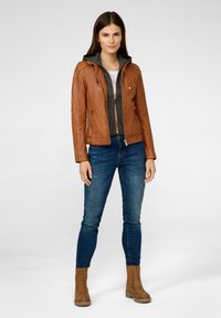 Bruine leren jas met een grijze hoodie, ritssluiting aan de voorkant, twee zijzakken, gecombineerd met blauwe jeans en tan kleurige enkellaarsjes.