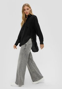 Zwarte, gestructureerde overhemd met een zak en zijsplitten, gecombineerd met grijze wijde jeans en witte sneakers, alles op een neutrale achtergrond.