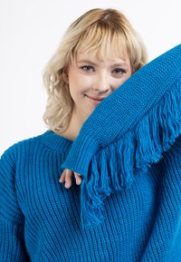Maglione blu lavorato a maglia con costine testurizzate, maniche oversize e dettagli di frangiatura ai polsi. Realizzato in morbido filato spesso.