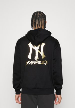Osoba z zaplecionymi włosami nosząca czarną bluzę z kapturem z dużym złotym logo New York Yankees i napisem na plecach.