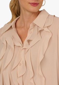 Pale pink blouse with ruffled detailing along the front and collar, featuring button closures and a textured fabric.