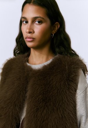 Brown fur vest with a soft, fluffy texture, worn over a light gray sweater, featuring a high collar and deep-cut armholes.