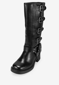 Bota de cuero negro hasta la rodilla con múltiples hebillas plateadas y un tacón grueso. Presenta una textura suave y una correa ajustable en el tobillo.