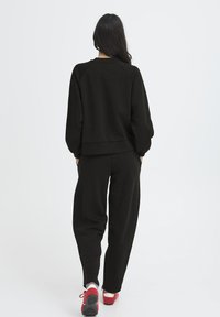 Melnas džemperis un saskaņotas bikses brīvā piegriezumā. Audumam ir gluda tekstūra; džemperim ir ribotas aproces un apakša. Sarkanas sporta kurpes.