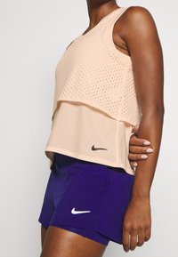 Canottiera leggera in rete pesca sovrapposta a uno strato interno solido, abbinata a pantaloni corti viola con logo Nike bianco.