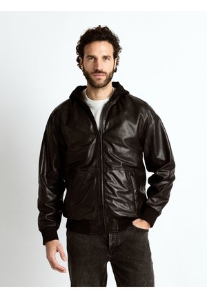 Mann mit lockigem Haar und Bart trägt eine schwarze Lederjacke mit Reißverschluss über einem weißen Hemd und posiert mit einer Hand in der Tasche vor schlichtem Hintergrund.