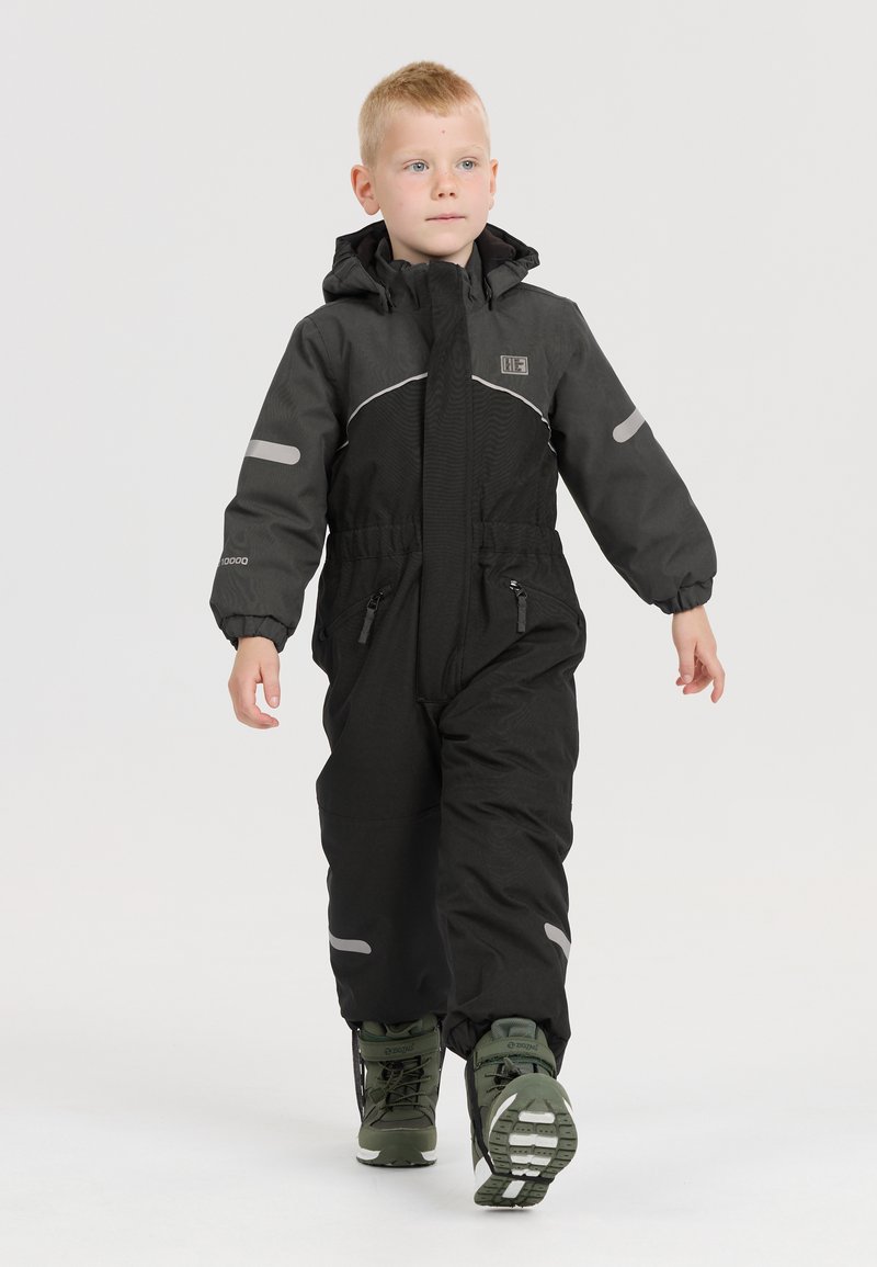 Combinaison de ski noire isolée, avec des accents réfléchissants et une fermeture éclair sur le devant, associée à des bottes d'hiver vertes, adaptée aux temps froids.