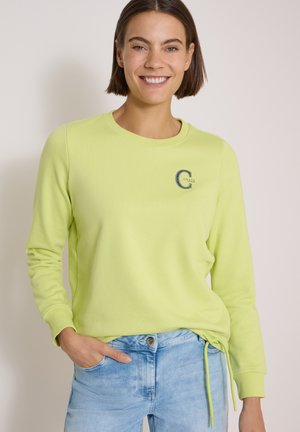 Jeune femme souriante, portant un haut à manches longues vert clair avec un petit logo sur la poitrine et un jean bleu clair, main dans la poche, fond neutre.