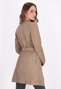 Beige ruskindsbæltefrakke med en tætsiddende silhuet, lange ærmer og en knælang kant. Har et rent bagside-design og minimal hardware.