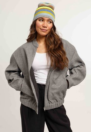 Donna che indossa una giacca grigia con zip, una canottiera bianca, pantaloni neri e un berretto di maglia a righe nei colori blu, giallo e grigio.