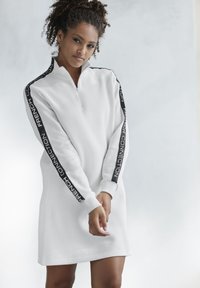 Weißes Fleece-Kleid mit Reißverschlusshals, ausgestattet mit schwarzem Text entlang der Ärmel. Gerippte Bündchen und lässiger Schnitt.