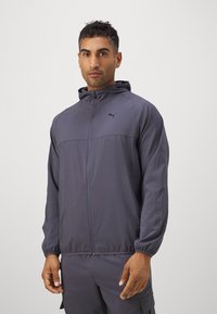 Puma hættetrøje i mørkegrå, med lynlås foran, elastiske manchetter og en glat tekstur. Logo accent på brystområdet.