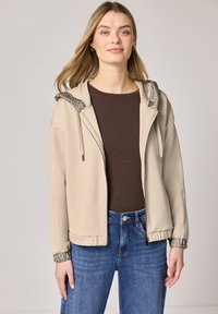 Kvinde iført beige hættejakke med lynlås og dyreprintkant, mørkebrun top og blå jeans, stående mod en ensartet lys baggrund.